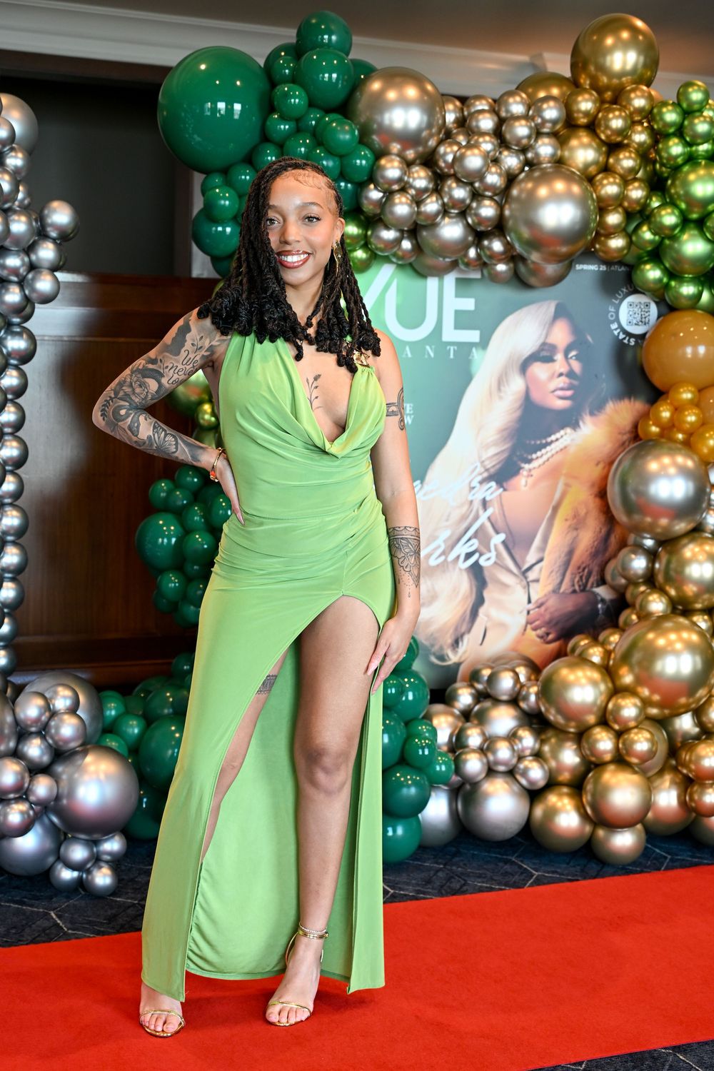 Model in lime green dress poses on red carpet with balloon arch backdrop at VUE magazine event.