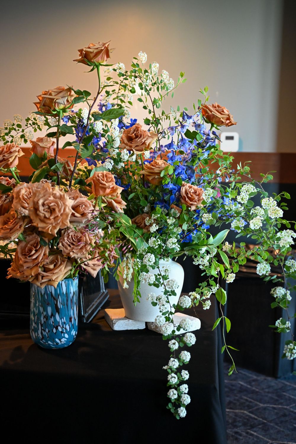 Elegant floral arrangement with copper-colored roses, white flowers, and blue blooms cascading over a dark surface.