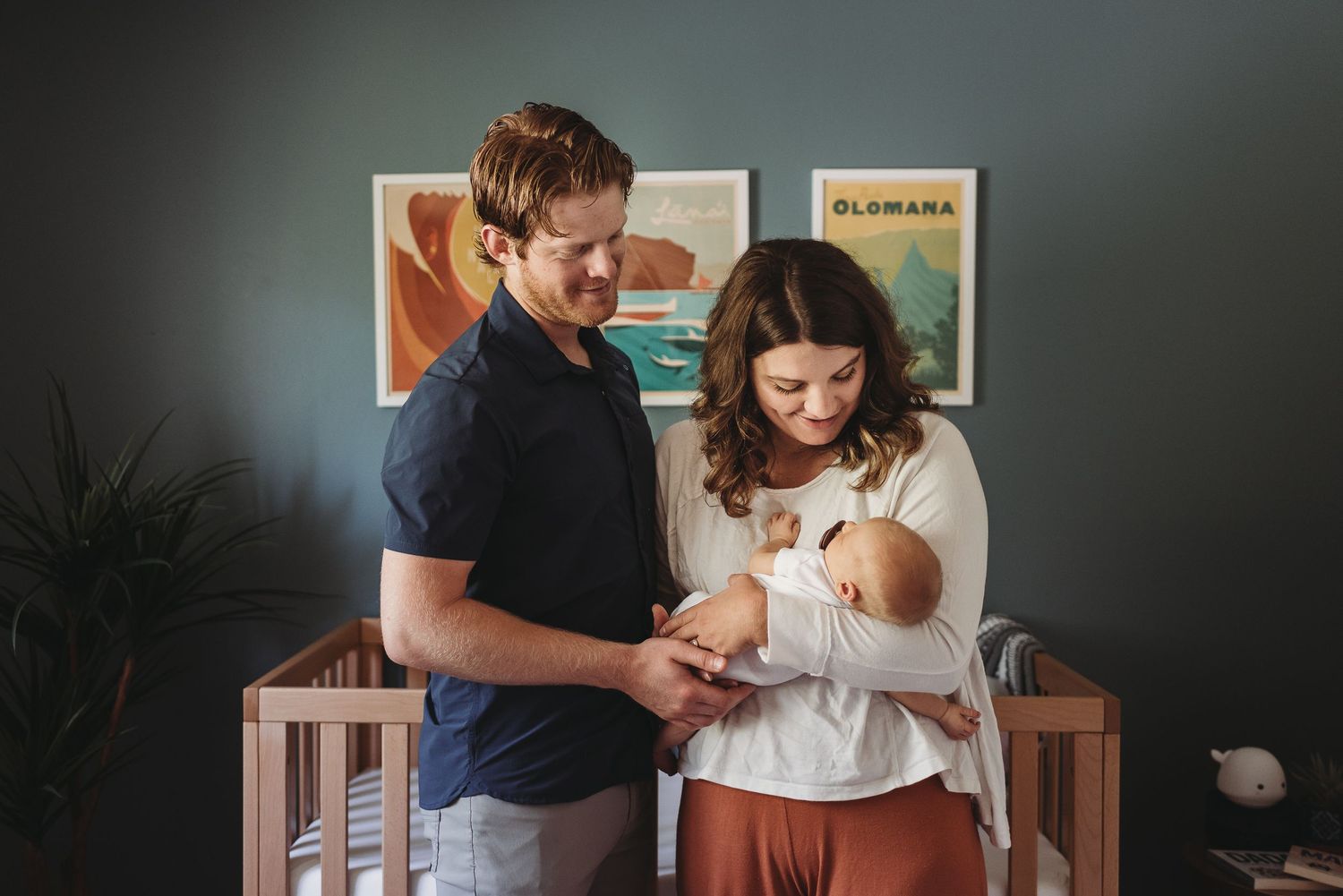 Parents in a dimly lit nursery room spending tender moments with their newborn baby in a wooden crib.