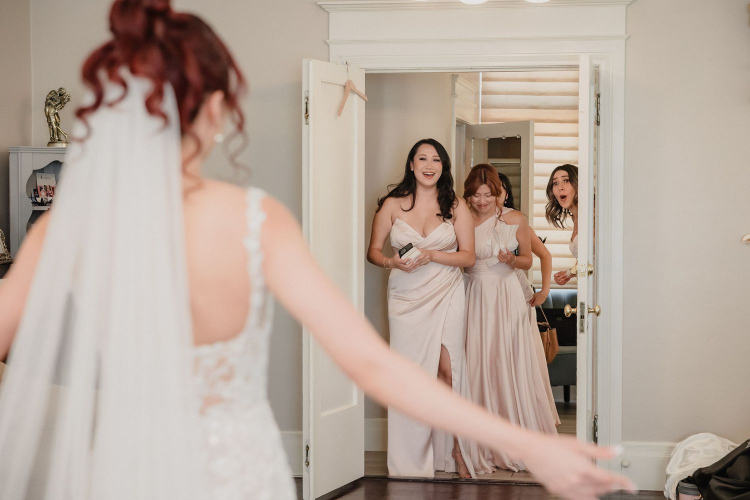 Bridesmaids in blush dresses react joyfully while watching wedding preparations in a mirror reflection.