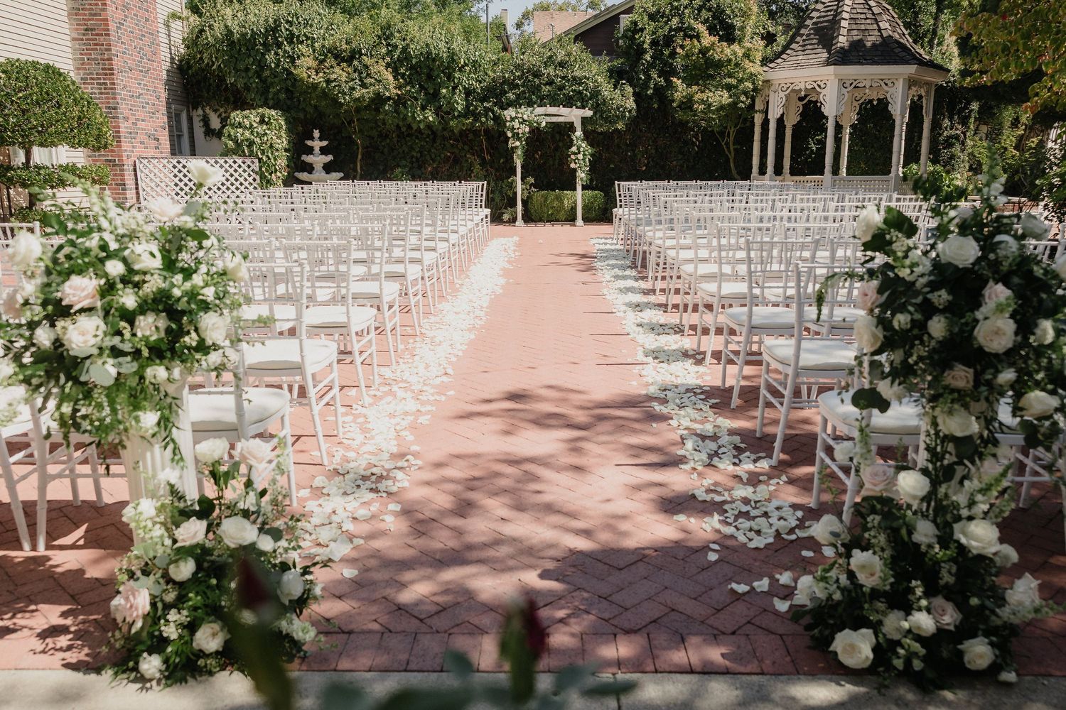 Outdoor wedding ceremony setup with white chairs and floral arch on brick patio.