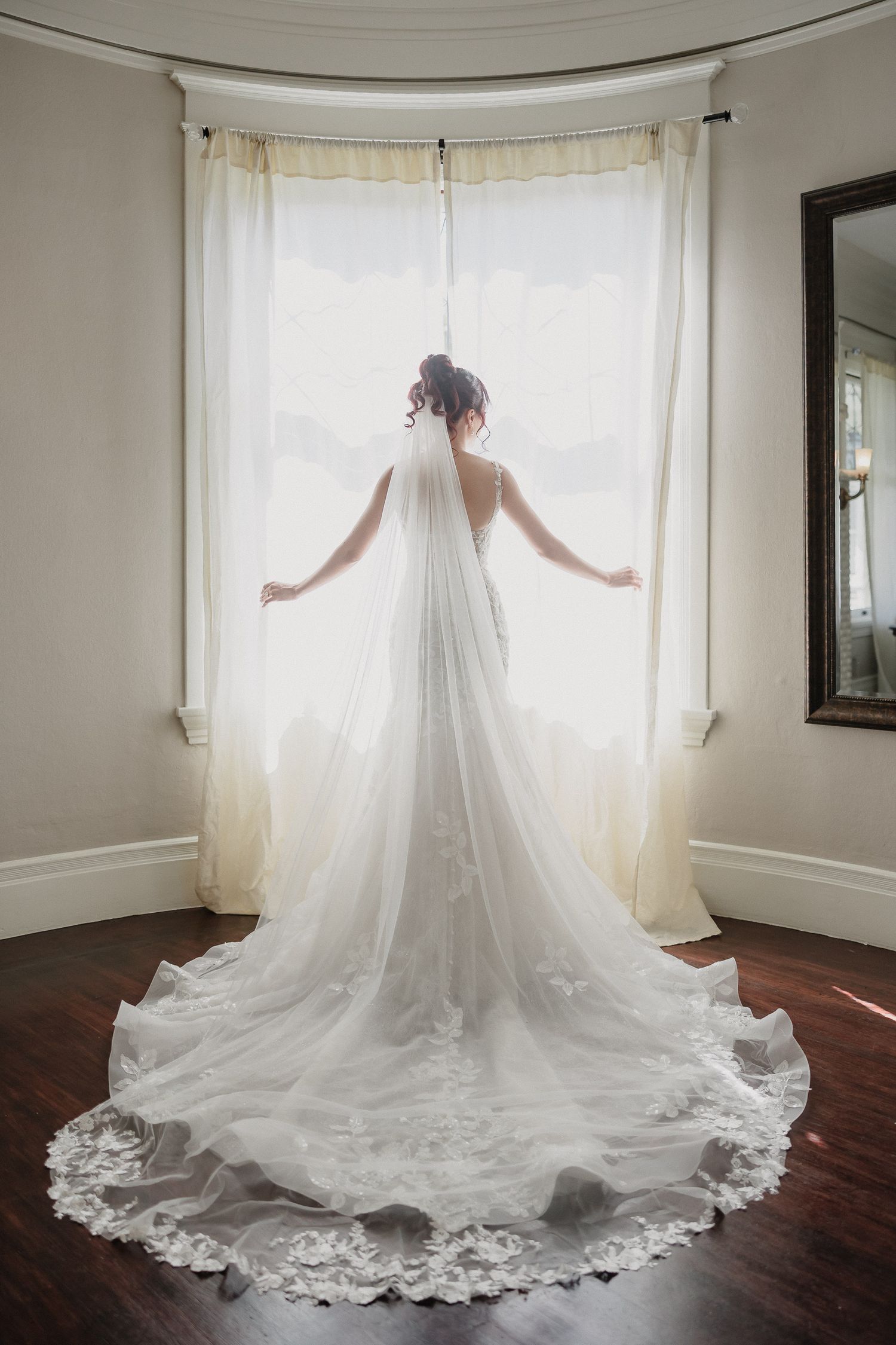 Silhouette of bride in flowing lace train wedding dress against bay window.