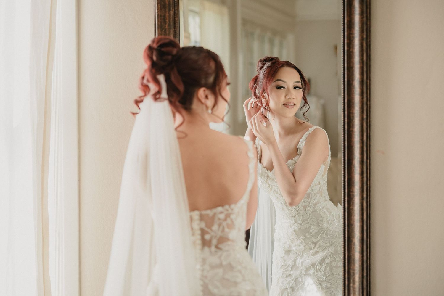 Bride in lace wedding gown admires her reflection in an elegant mirror during wedding preparations.