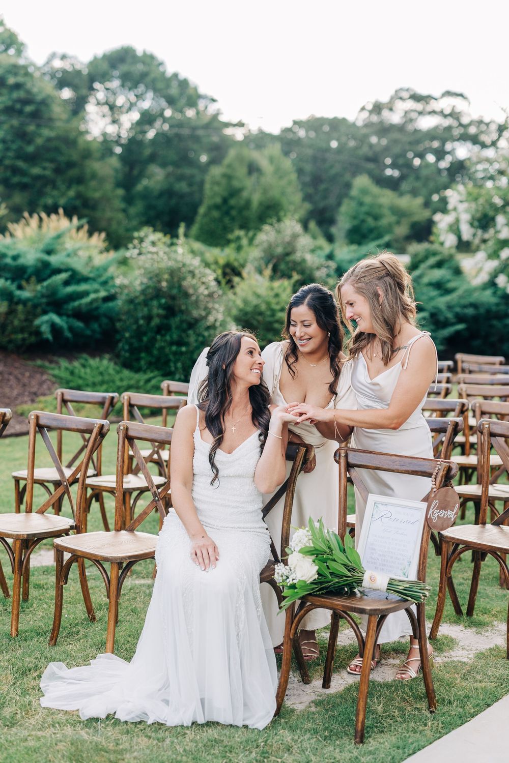 Celebration of joy between women in white dresses sitting on wooden chairs at an outdoor venue between trees and bushes.