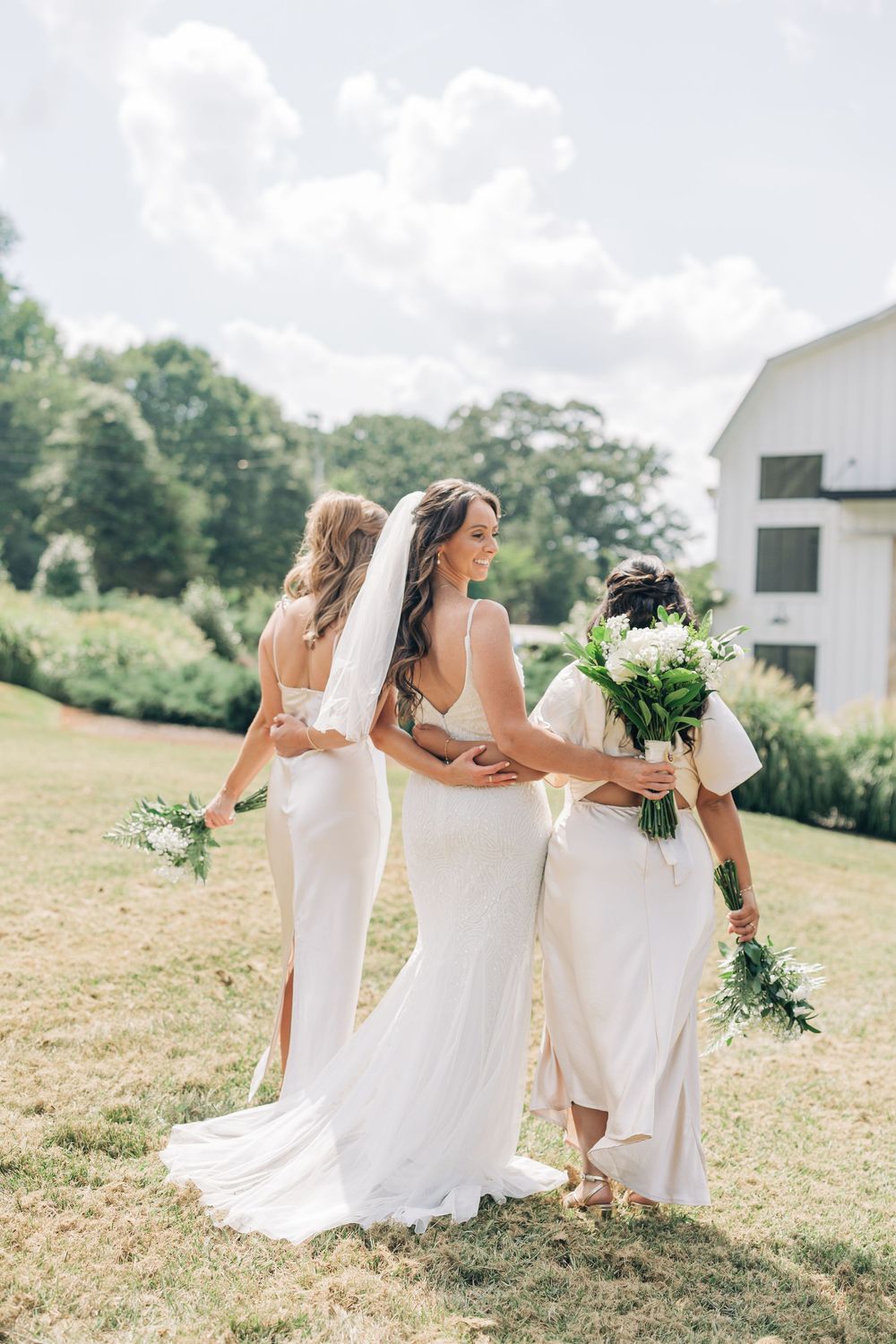 Three people in white dresses hold bouquets while posing in a scenic outdoor field on a sunny summer day.