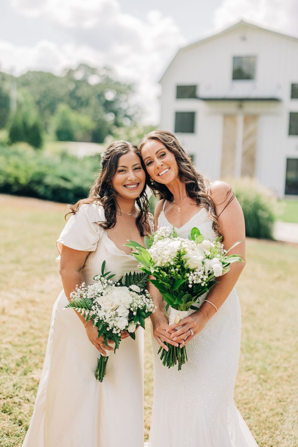 Two people in white dresses hold beautiful white and green floral bouquets while posing together on a lawn near a house.
