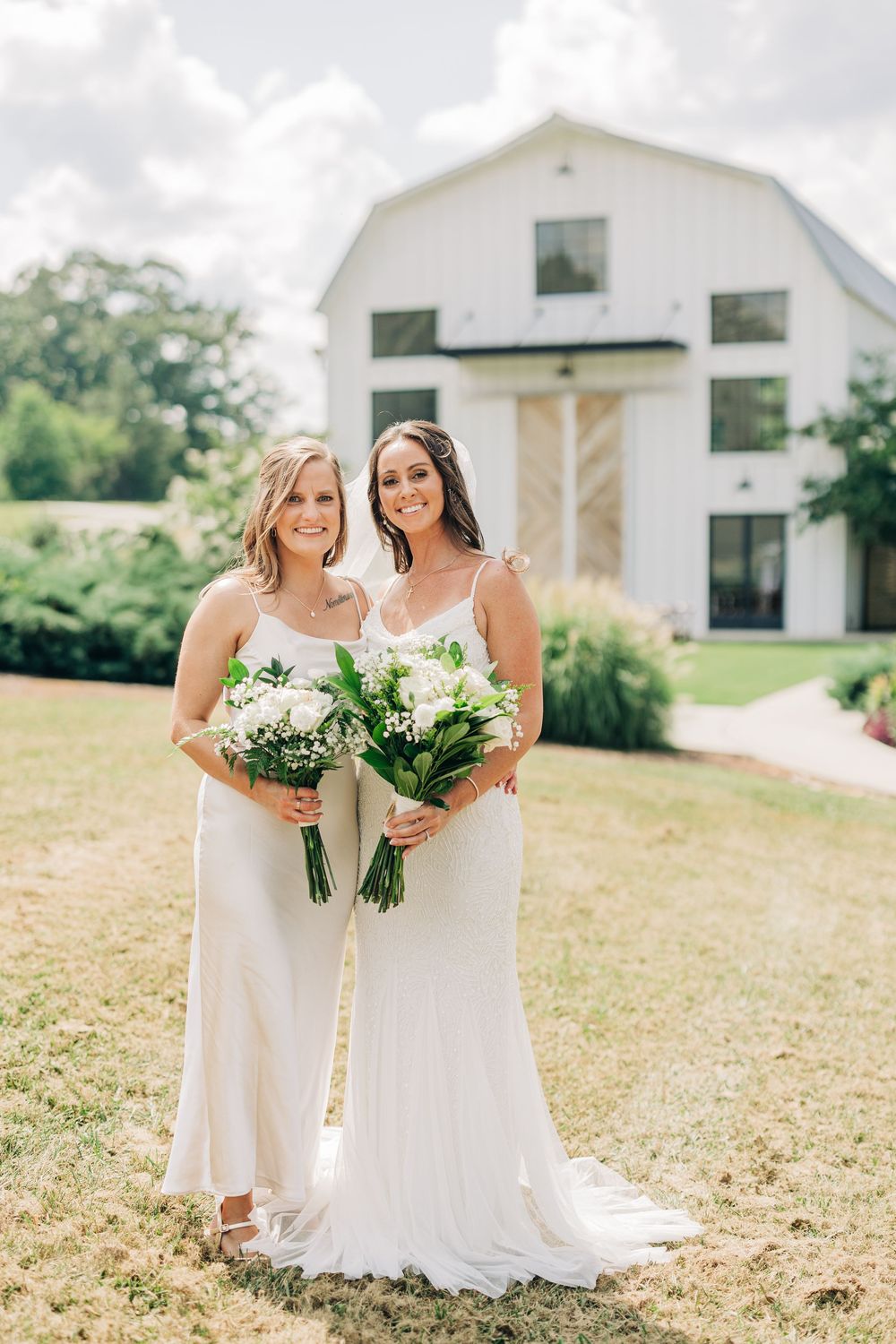 Two people in white dresses share a joyful moment together outdoors near a white barn on a sunny day.