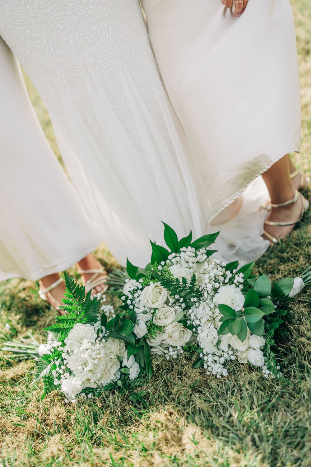 Close-up of a white wedding dress hem against grass with an elegant white and green floral bouquet arrangement.