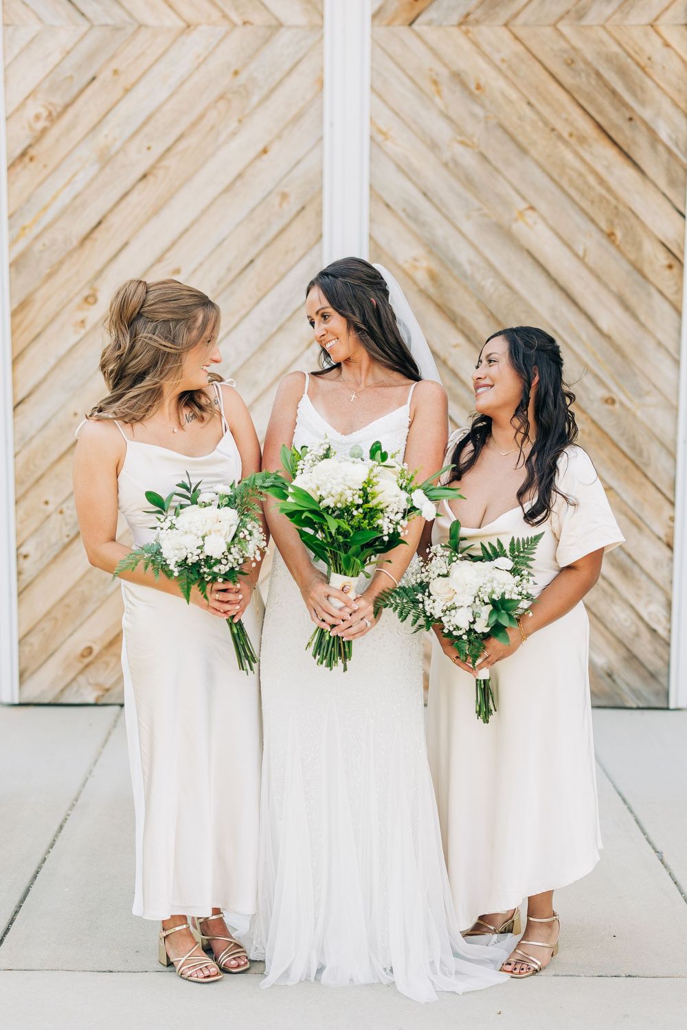 Bridesmaids in flowing white dresses holding elegant white and green bouquets pose together against a chevron wall pattern.