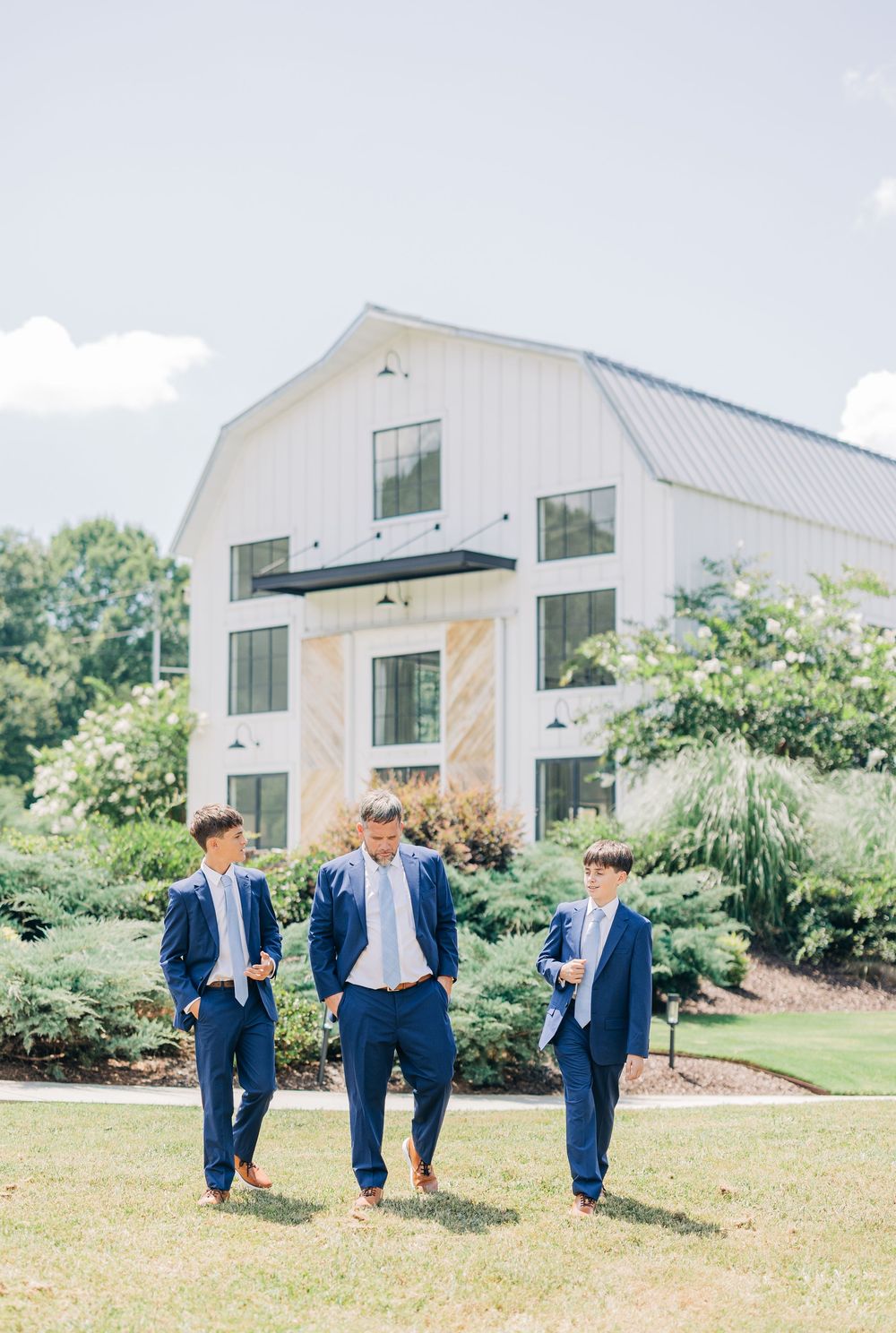 A group in navy blue suits playfully kicks and poses on a grassy lawn outside a modern white barn building.