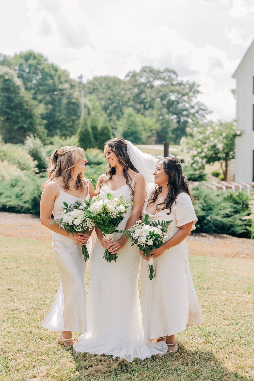 Four bridesmaids in white dresses stand together holding white floral bouquets in an outdoor garden setting.