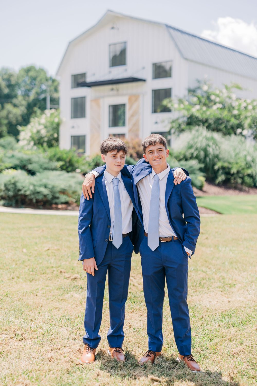 Young groomsmen in navy blue suits with light blue ties pose playfully on a sunny lawn in front of a white building.