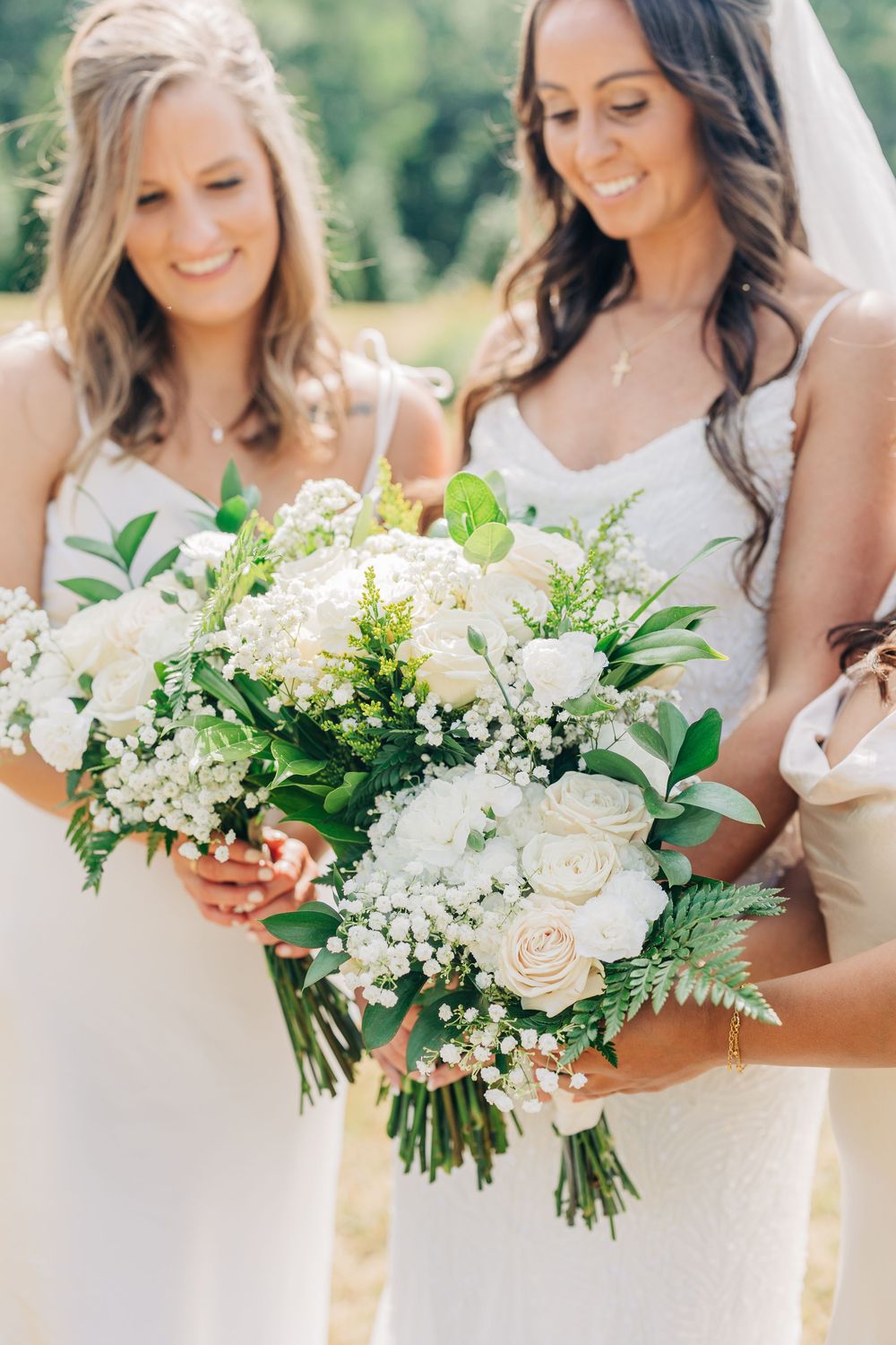A row of wedding bouquets featuring white roses, baby's breath and greenery held against white dresses in natural light.