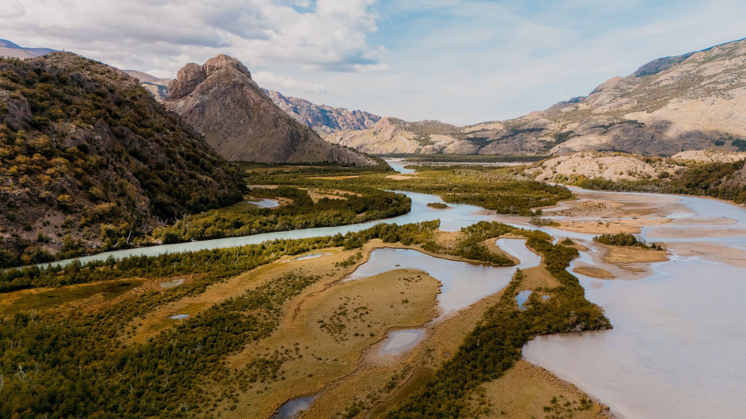 Meandering river flows through a scenic mountain valley with rugged peaks and golden terrain under cloudy skies.
