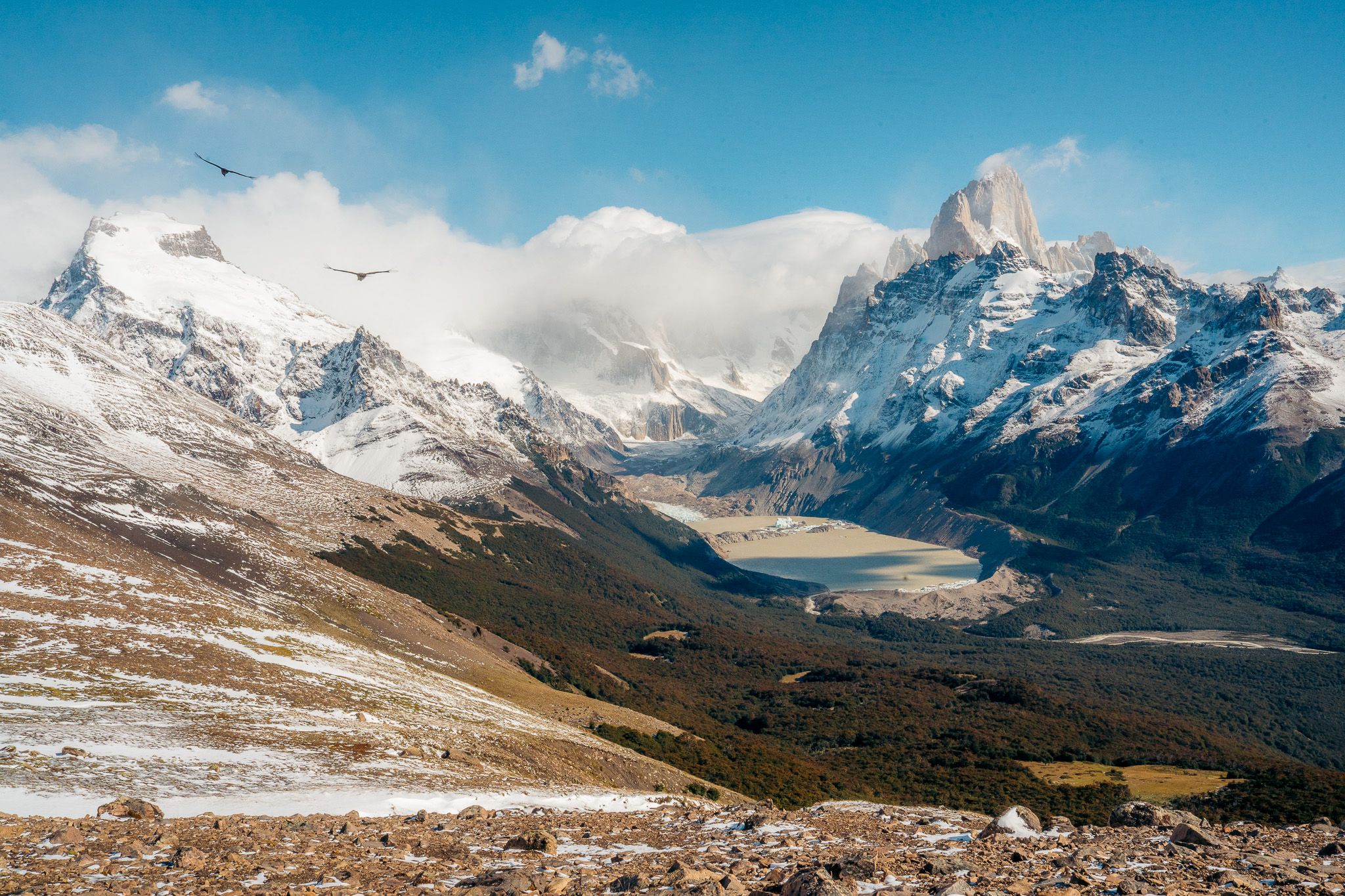 A panoramic mountain landscape showing snow-capped peaks, valleys, and rugged terrain under a blue sky.