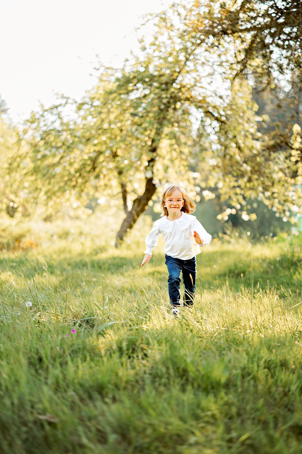 A joyful young girl in a white top and jeans runs through a sunny green meadow with blooming trees in the background.