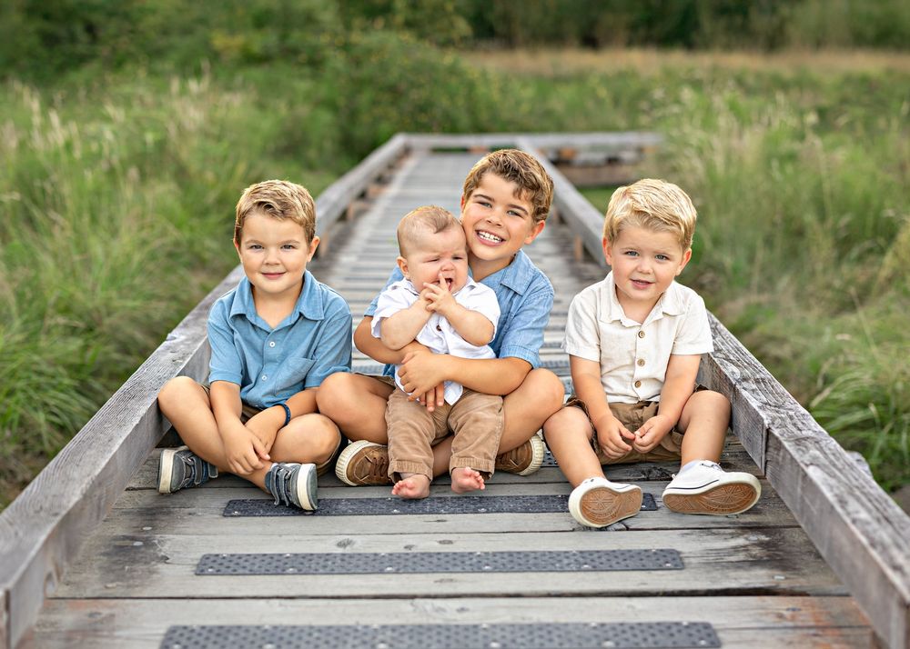 Four young boys sitting together on a wooden bridge outdoors, smiling for a family portrait in a green field.