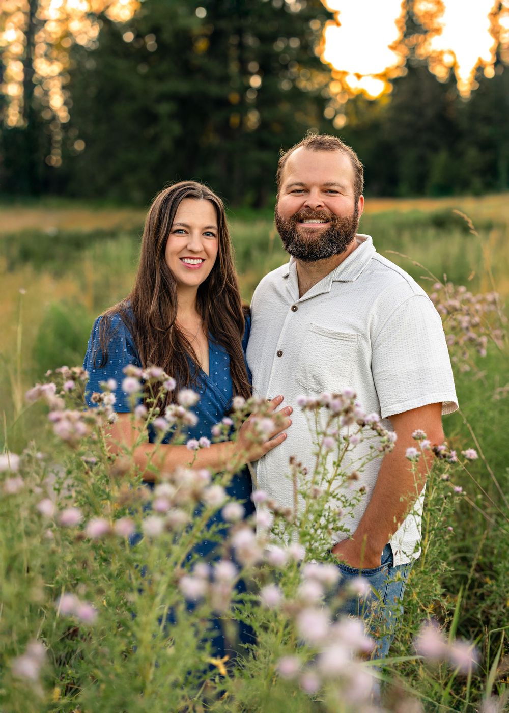 A smiling couple stands together in a wildflower meadow at golden hour with tall pine trees in the background.