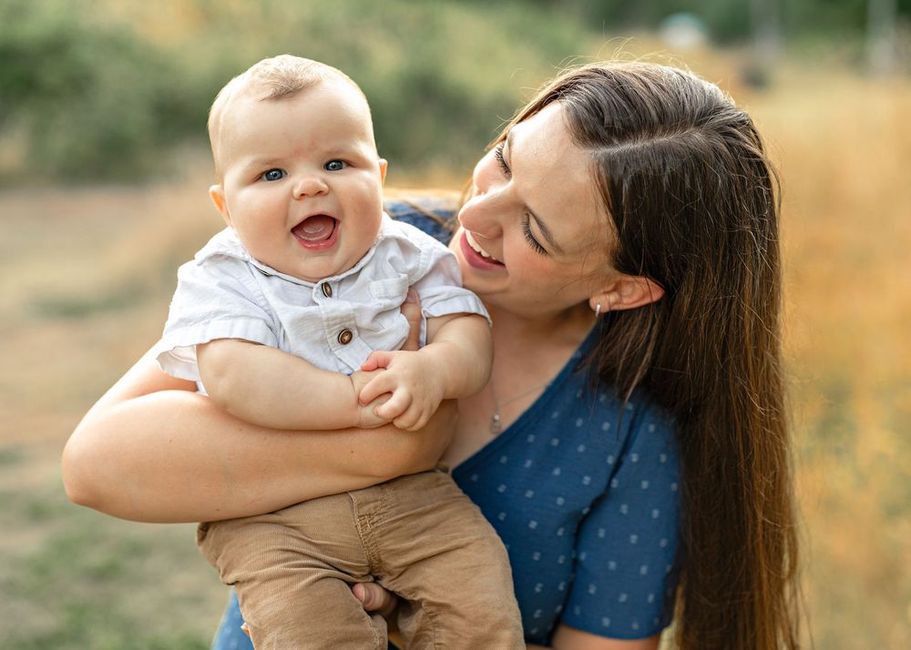 A smiling mother holds her happy laughing baby boy outdoors in a sun-lit field during a family photo session.