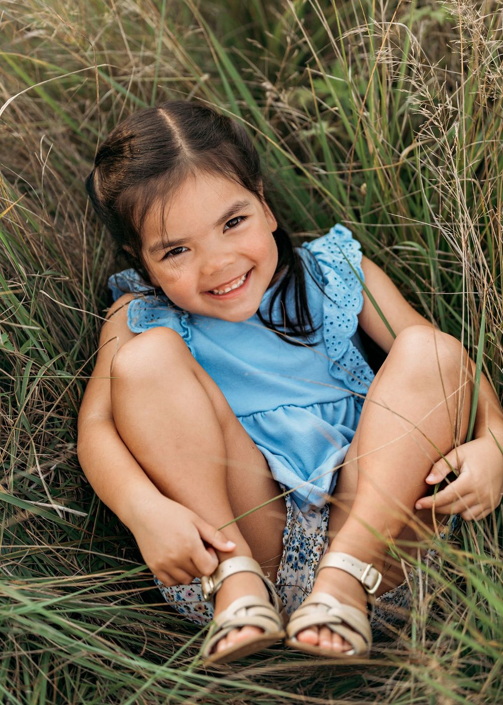 A child in a blue dress sitting in tall grass with sandals.