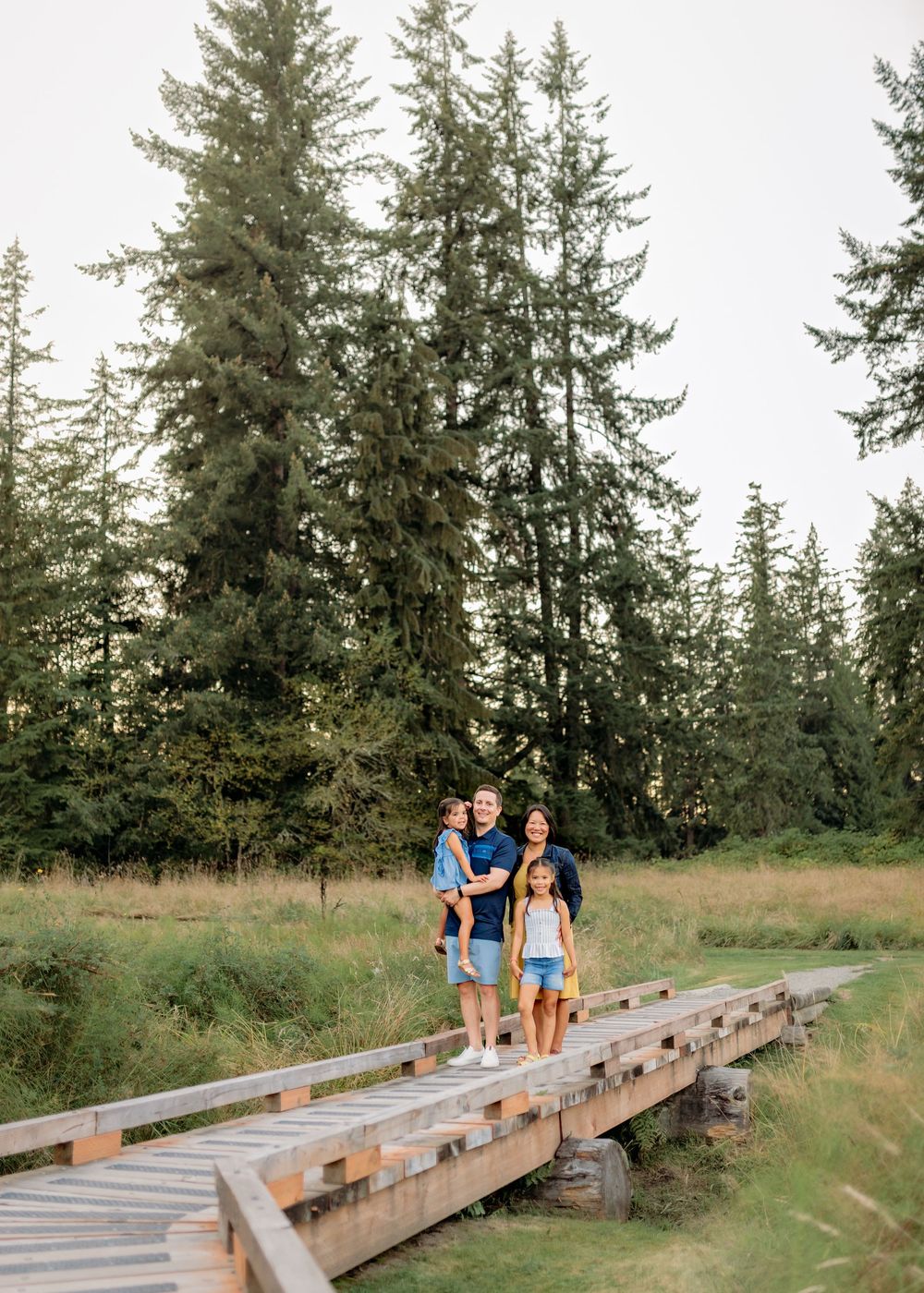 A family walking together on a wooden boardwalk surrounded by tall evergreen trees.