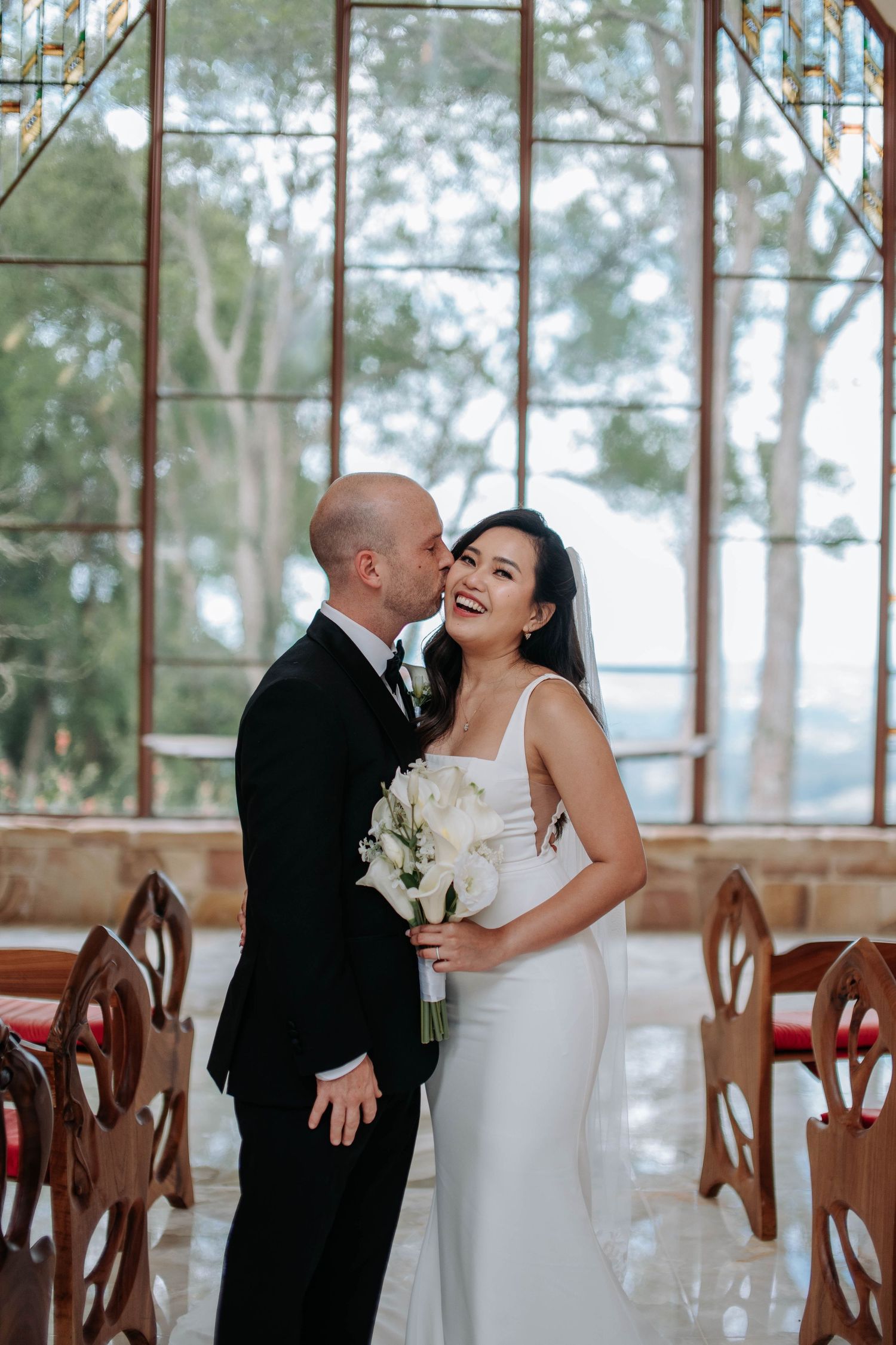 A romantic wedding moment captured in a glass-walled venue with wooden chairs and natural light filtering through trees.