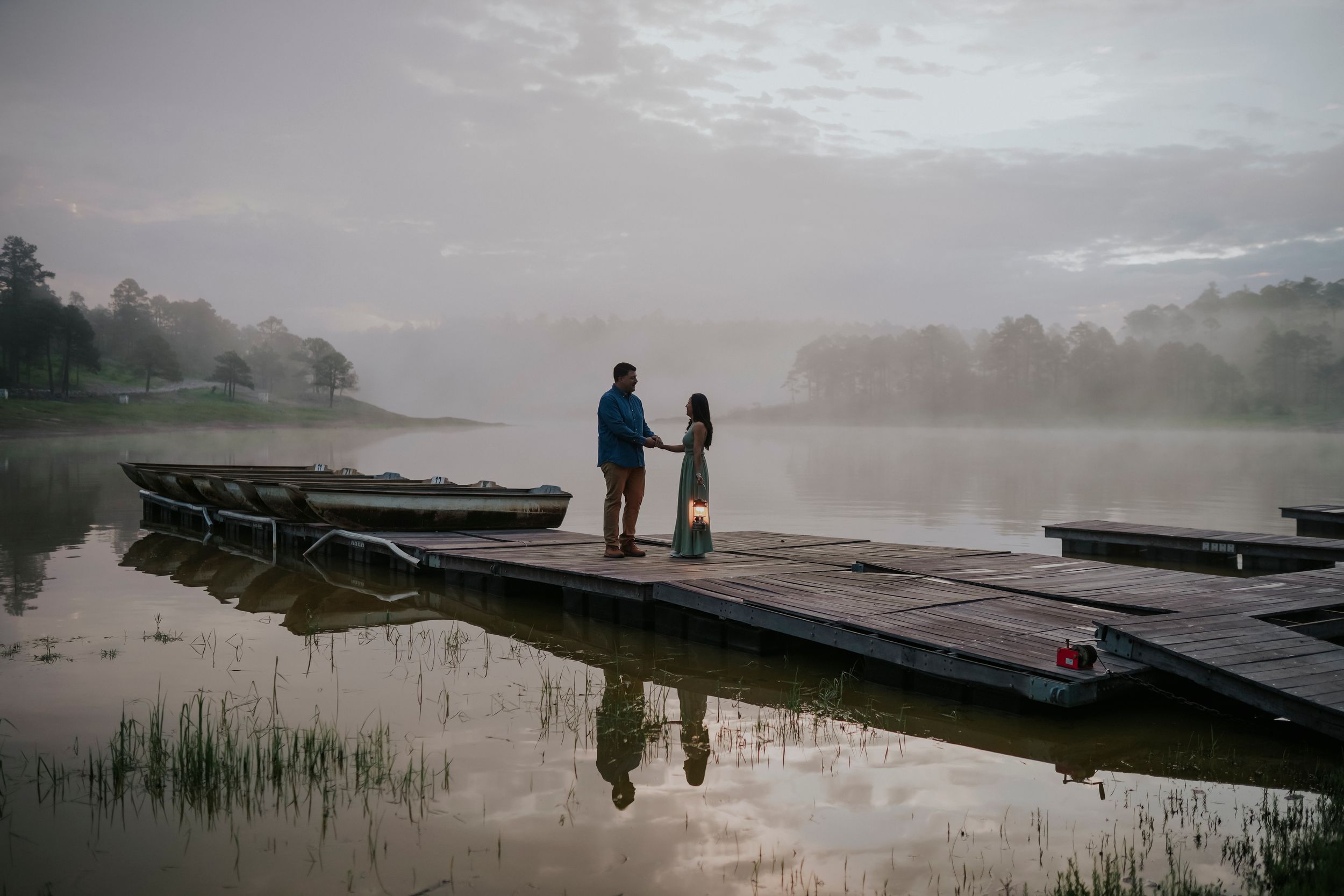 Two silhouettes stand together on a misty lakeside dock at dawn while fog rises over the tranquil water.