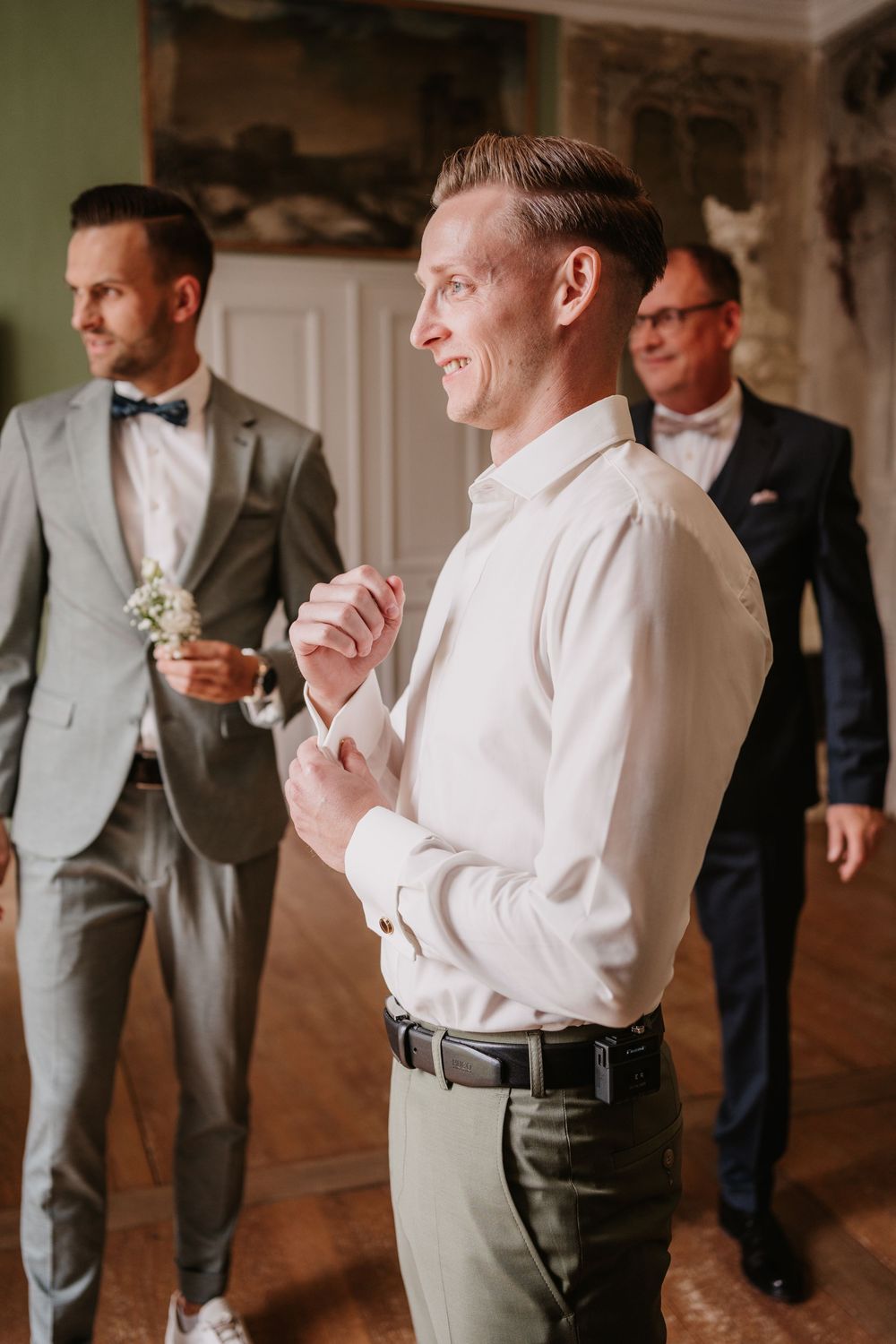 Groomsmen getting ready in formal attire at an elegant indoor venue with white walls and warm lighting.