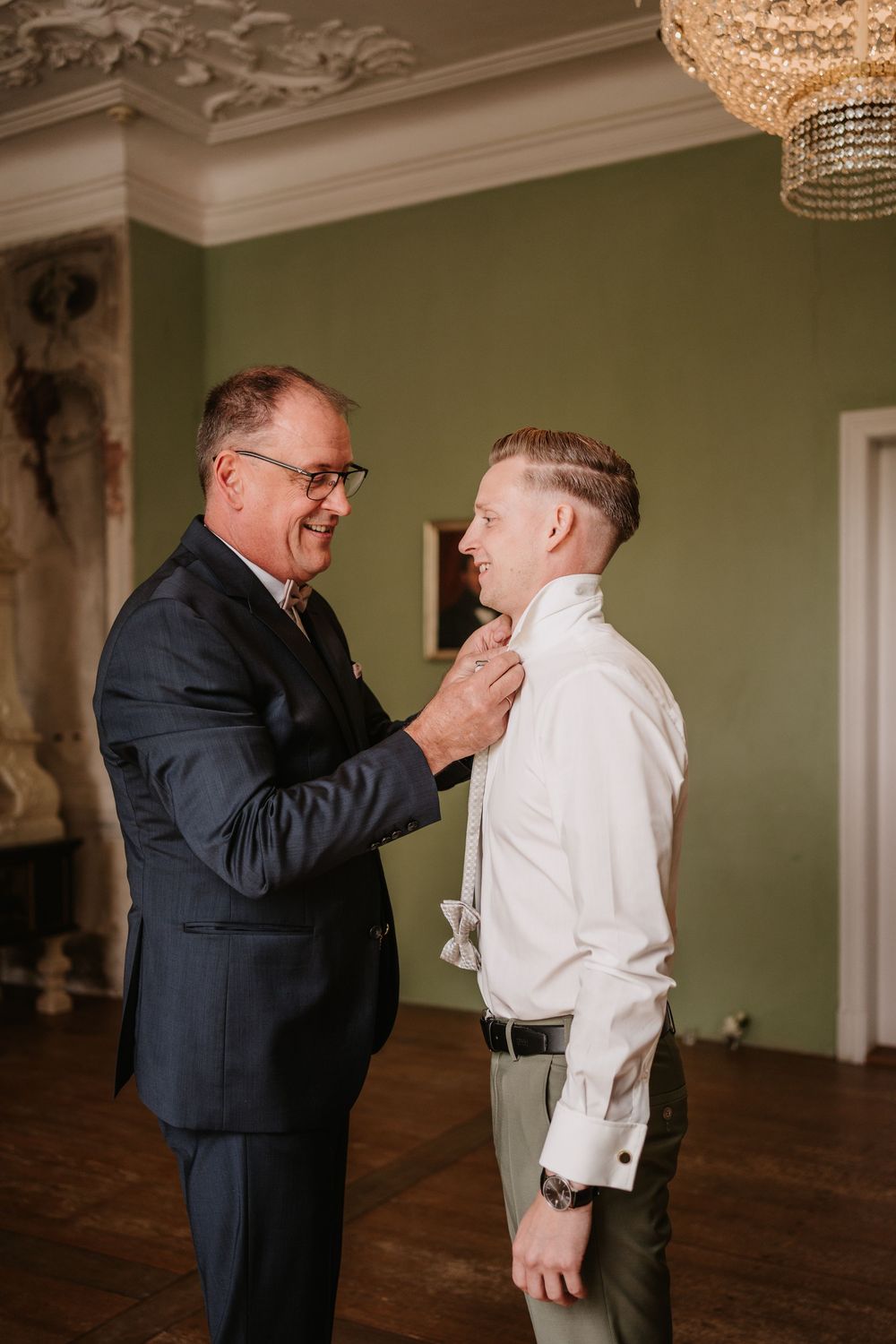 Two men stand in an elegant room with green walls as one helps adjust the other's tie before a formal event.