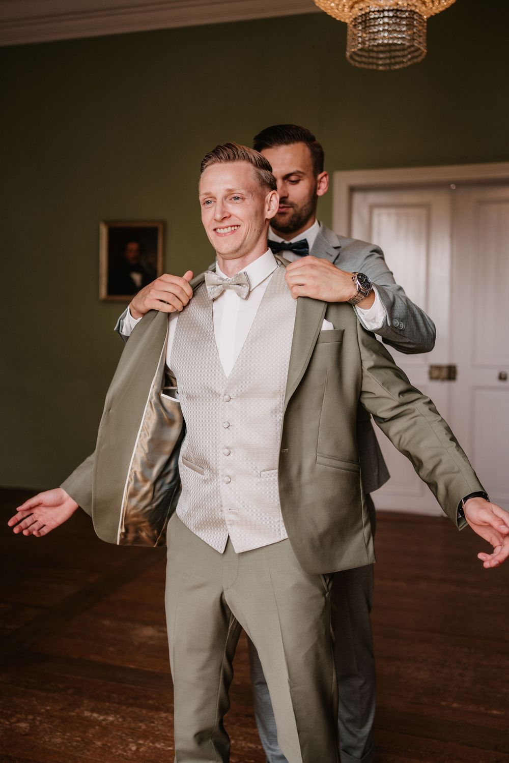 Wedding party members helping with formal attire preparations in a room with a crystal chandelier.