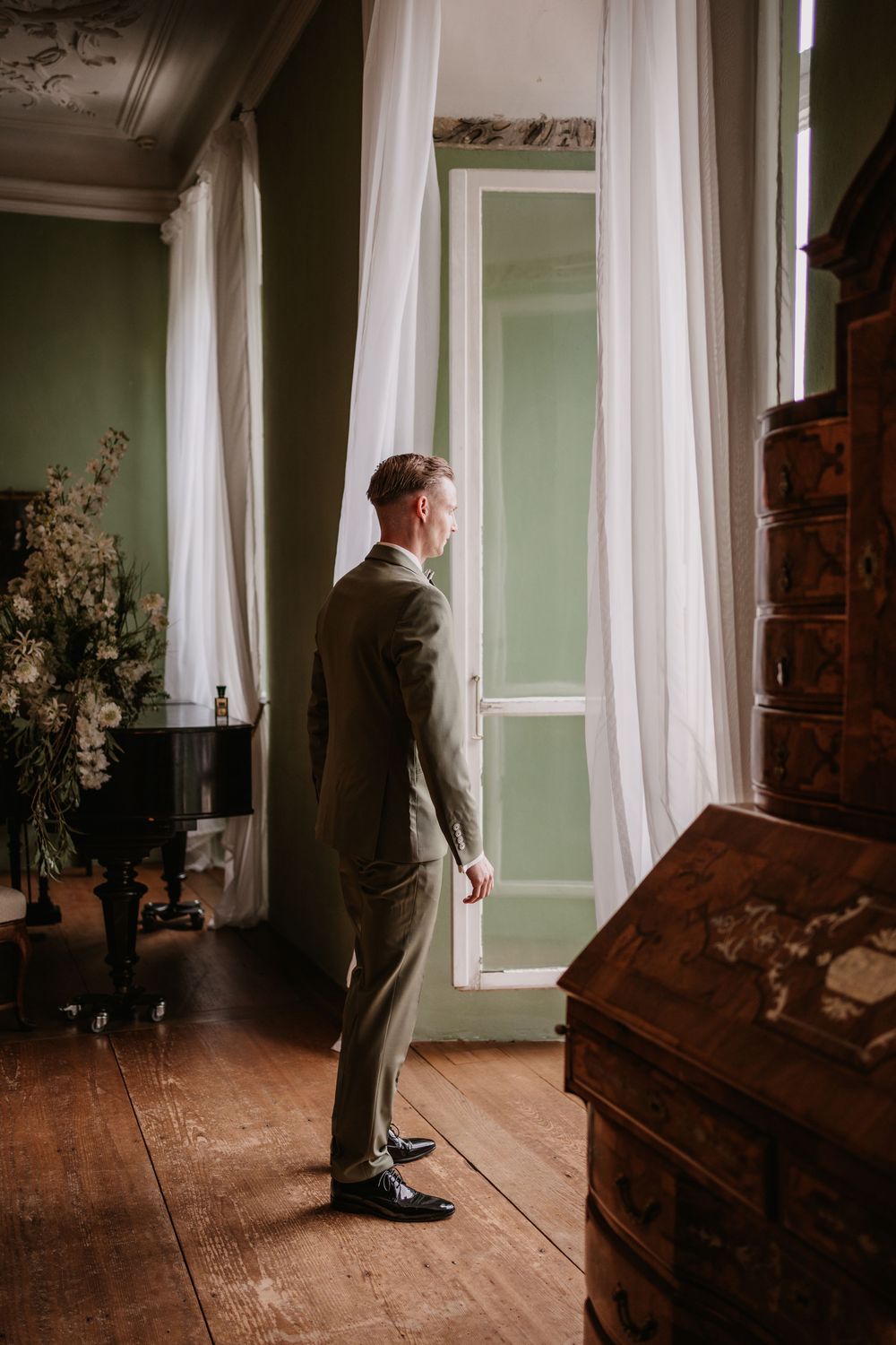 A person in a gray suit stands near a window in a room with wooden floors and a vintage bureau.