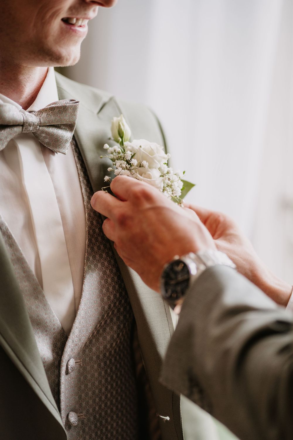 Close-up of a groom's formal attire showing a bow tie, textured vest, and delicate white floral boutonniere.
