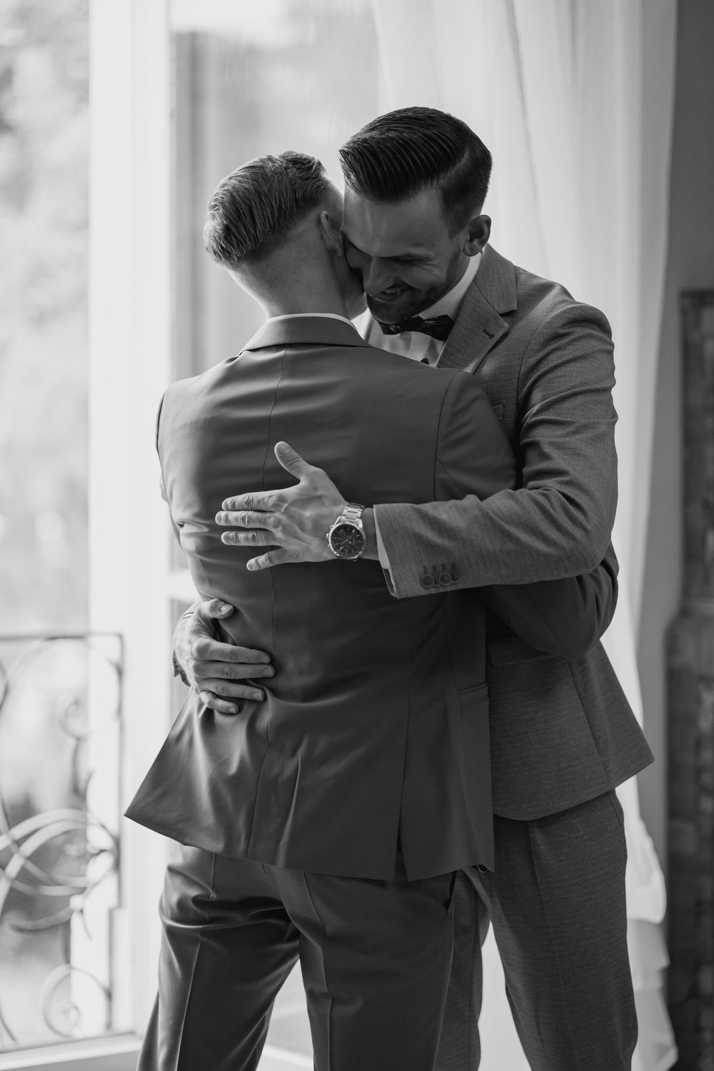 Two men in grey suits share an intimate embrace near a window in a black and white photograph.