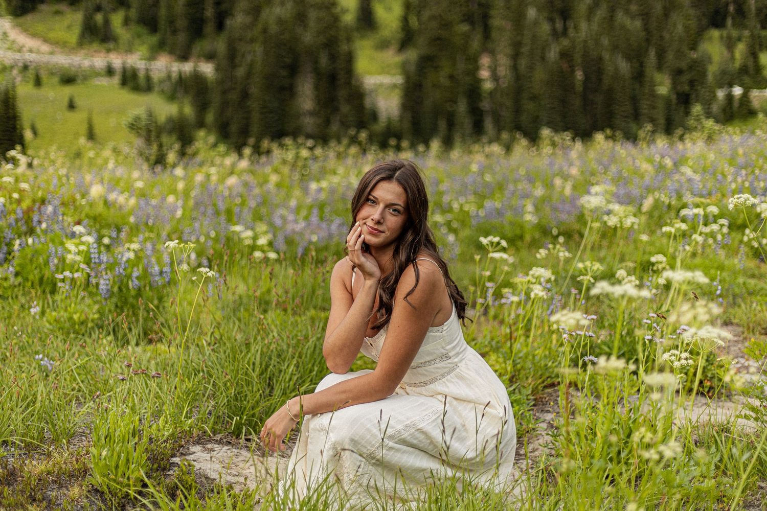 A figure in a flowing white dress poses gracefully on a rocky outcrop surrounded by mountain wildflowers and evergreens.