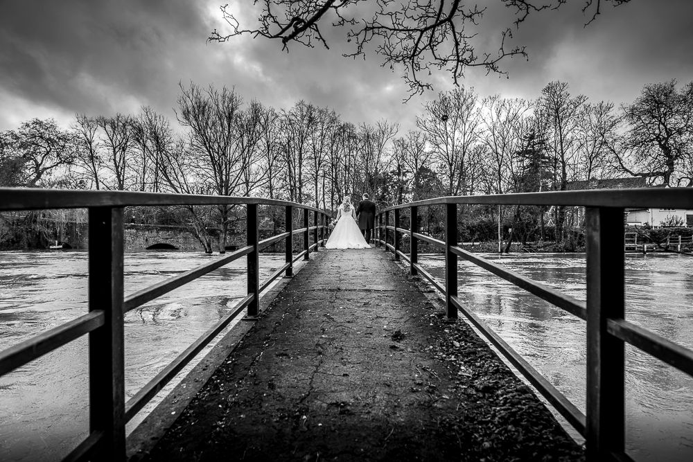 Dramatic black and white photograph of a long footbridge over water surrounded by bare trees.