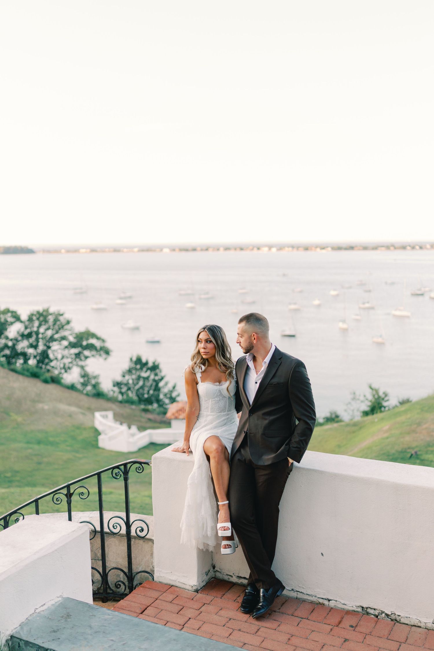 Bride and groom pose at Vanderbilt Museum overlooking a harbor with boats at sunset.