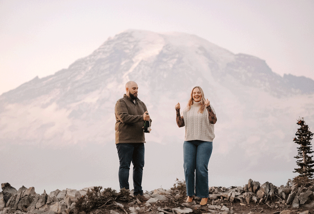Couple celebrating engagement on rocky mountain summit with snow-capped Mount Rainier in background.