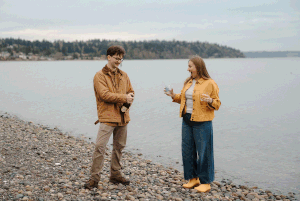 Two people in yellow jackets chat on a rocky beach shore with calm water and trees in the background.