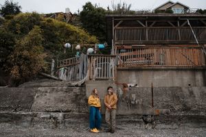 Two people in yellow jackets stand against a weathered concrete wall near a wooden deck with colorful buoys.