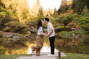 Couple sharing a romantic kiss on a wooden dock beside a serene Japanese garden pond surrounded by lush trees.