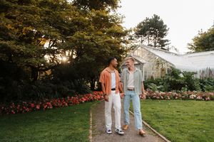 Two men walking together on a garden path surrounded by lush greenery and red flowers near a greenhouse.
