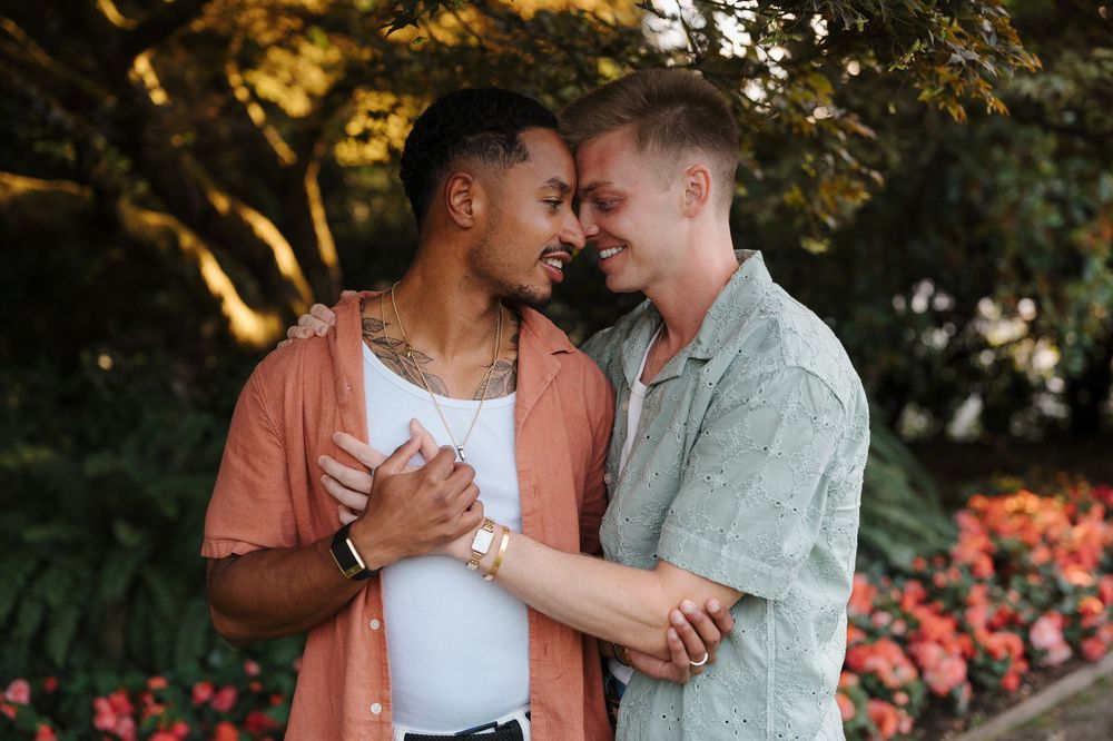 Two men sharing a romantic moment outdoors, smiling and embracing near colorful flowers and lush green trees.