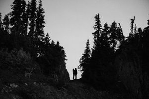 Silhouette of two people standing on a rocky ridge surrounded by tall pine trees in a black and white landscape photo.