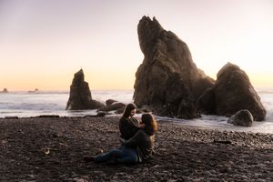 Couple embracing on a rocky black sand beach at sunset with dramatic sea stacks rising from the ocean.