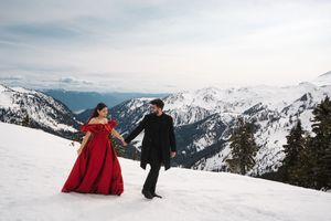 Romantic winter engagement photoshoot with couple sitting in snow against majestic mountain backdrop.