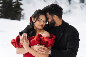 A couple in formal attire embraces romantically against a snowy winter backdrop with pine trees.