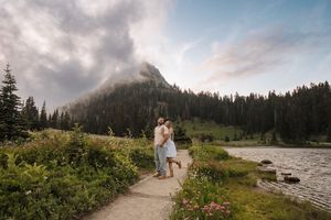 A scenic mountain hiking trail surrounded by wildflowers and evergreen trees with dramatic cloudy skies overhead.