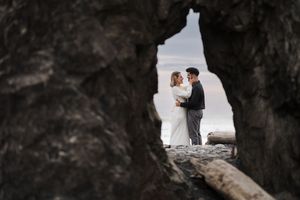 A romantic wedding couple stands framed within a rocky cave opening overlooking a misty landscape.