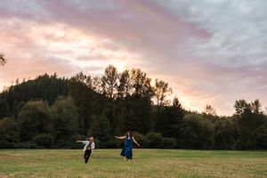 People walking on a green field at sunset with trees silhouetted against a dramatic pink and purple sky.
