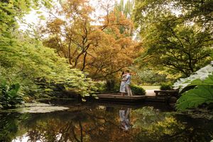 A couple stands on a wooden bridge over a reflective pond surrounded by lush autumn foliage in a garden setting.